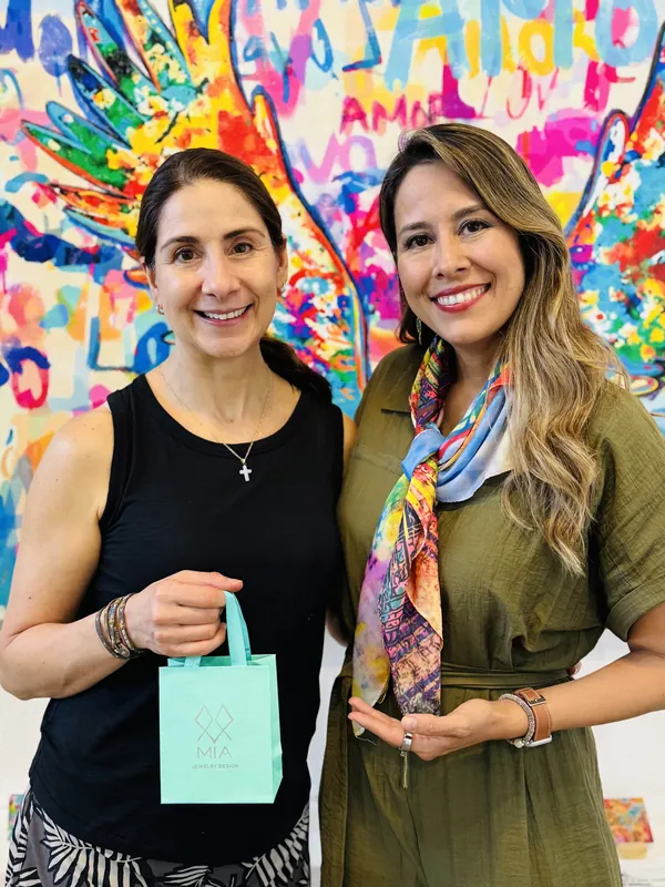 Two friends at the colorful angel-wings mural, one holding the aqua MIA gift bag, the other wearing her silver cross necklace