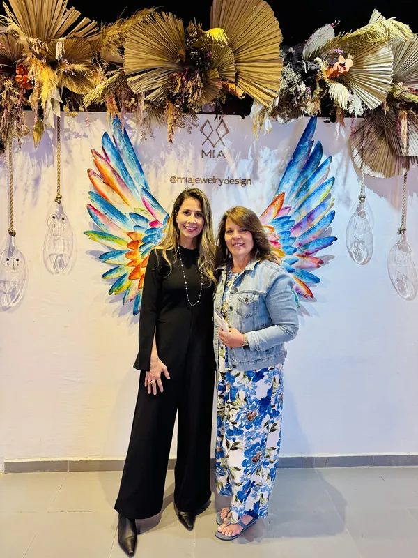 Imelda wearing her pearl necklace beside a client in a denim jacket at the angel-wings backdrop