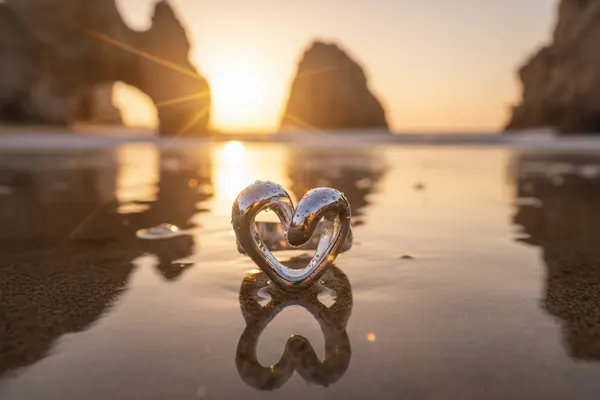 Protect My Love silver heart ring standing on wet sand with its reflection, sea arch rocks at sunset