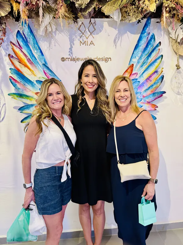 Imelda in black between two smiling clients, one carrying the aqua MIA gift bag, in front of the painted angel-wings backdrop